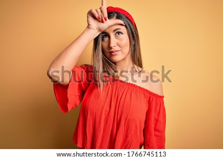 Young beautiful woman colorful summer style over yellow isolated background making fun of people with fingers on forehead doing loser gesture mocking and insulting.