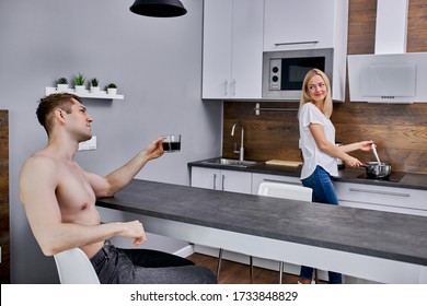 young beautiful woman cooking breakfast in the morning, handsome shirtless guy sit making compliments his wife while drinking coffee - Powered by Shutterstock