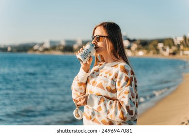 Young beautiful woman in cozy sweater and sunglasses enjoying hot coffee from thermo mug while relaxing on winter seaside sand beach. Cute attractive girl drinks tea taking sunbathe near ocean. - Powered by Shutterstock