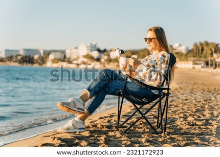Young beautiful woman in cozy sweater and sunglasses filling cup of coffee from thermos while relaxing on winter seaside sand beach. Cute attractive girl enjoying hot tea taking sunbathe near ocean.