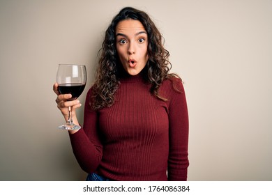 Young beautiful woman with curly hair drinking glass of red wine over white background scared in shock with a surprise face, afraid and excited with fear expression
