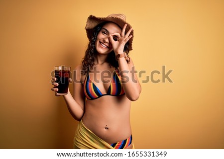 Young beautiful woman with curly hair on vacation wearing bikini drinking cola beverage with happy face smiling doing ok sign with hand on eye looking through fingers