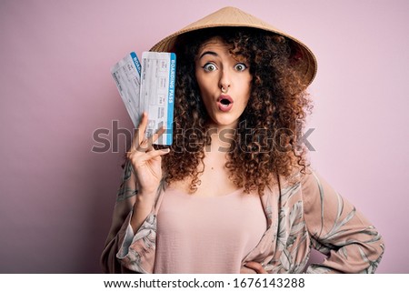 Young beautiful woman with curly hair and piercing wearing asian hat holding boarding pass scared in shock with a surprise face, afraid and excited with fear expression