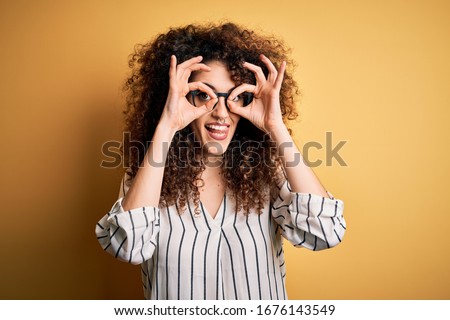 Young beautiful woman with curly hair and piercing wearing striped shirt and glasses doing ok gesture like binoculars sticking tongue out, eyes looking through fingers. Crazy expression.