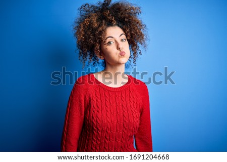 Young beautiful woman with curly hair and piercing wearing casual red sweater puffing cheeks with funny face. Mouth inflated with air, crazy expression.