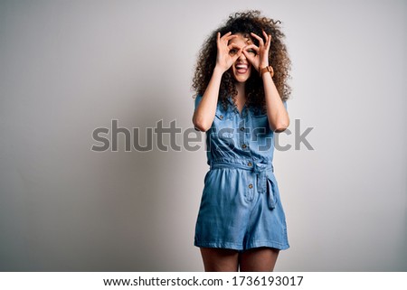 Young beautiful woman with curly hair and piercing wearing casual denim dress doing ok gesture like binoculars sticking tongue out, eyes looking through fingers. Crazy expression.
