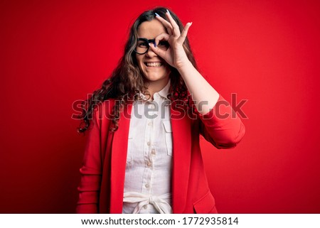 Young beautiful woman with curly hair wearing jacket and glasses over red background doing ok gesture with hand smiling, eye looking through fingers with happy face.