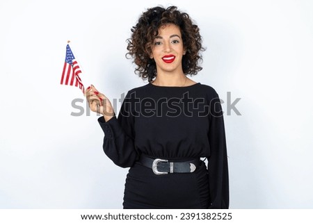 Young beautiful woman with curly hair wearing black dress and holding and American USA flag and smiling over white studio background. 