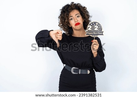 Young beautiful woman with curly hair wearing black dress over white studio background holding a Happy New year 2024 banner and holding thumb down
