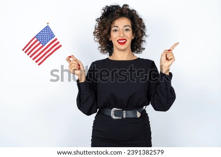 Young beautiful woman with curly hair wearing black dress and holding and American USA flag and pointing aside over white studio background. 