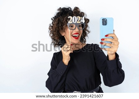 Young beautiful woman with curly hair wearing black dress, holding happy new year 2024 glasses and making a video call to friends and family with her smartphone .