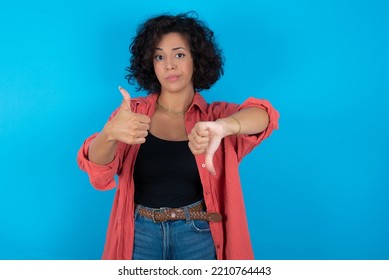 Young Beautiful Woman With Curly Short Hair Wearing Red Overshirt Over Blue Wall Feeling Unsure Making Good Bad Sign. Displeased And Unimpressed.