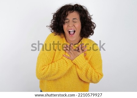 young beautiful woman with curly short hair wearing yellow sweater over white wall expresses happiness, laughs pleasantly, keeps hands on heart