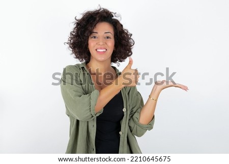 young beautiful woman with curly short hair wearing green overshirt over white wall Showing palm hand and doing ok gesture with thumbs up, smiling happy and cheerful.