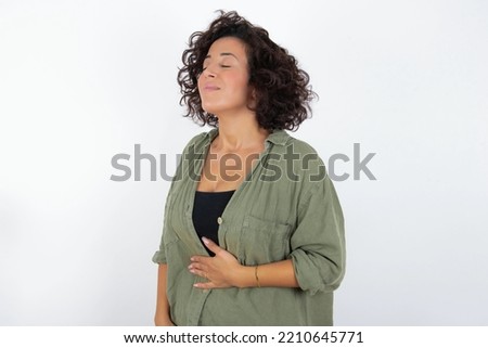 young beautiful woman with curly short hair wearing green overshirt over white wall touches tummy, smiles gently, eating and satisfaction concept.