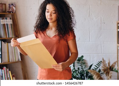 Young beautiful woman with dark curly hair in T-shirt dreamily opening envelope with exam results in hands with bookshelf on background at cozy home