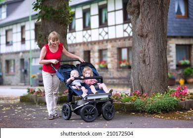 Young beautiful woman with a double jogging stroller with two kids, baby boy and little toddler girl, brother and sister, walking in a beautiful German village with traditional old houses