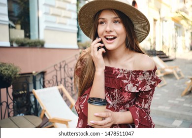 Young Beautiful Woman In Dress And Straw Hat Holding In Hand Cup Of Coffee To Go And Talking On Celphone Happily Looking Aside On Cozy City Street