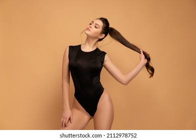 Young beautiful woman dressed black bodysuit standing studio isolated on color background holding hair ponytail looking away. Caucasian female in black swimwear orange background.