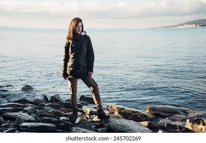 Young Beautiful Woman Dressed In A Coat And Cargo Pants Standing On Rocky Shore Near The Sea