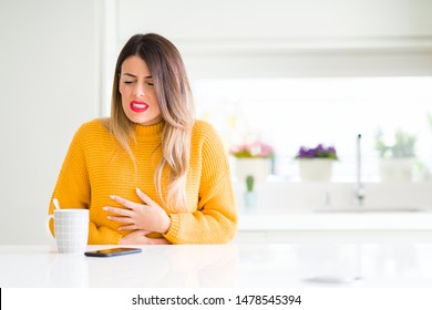 Young beautiful woman drinking a cup of coffee at home with hand on stomach because nausea, painful disease feeling unwell. Ache concept.