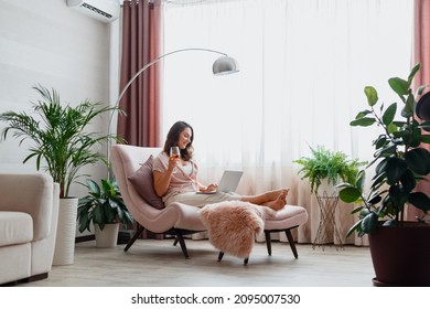 Young beautiful woman drinking rose wine and using laptop while sitting on comfortable pink armchair in home interior near window. Relax woman watching movie on laptop or make online purchases.