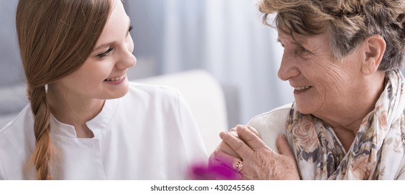 Young beautiful woman in a duster and her lovely elderly patient in a nursing home. Both women talking and smiling together