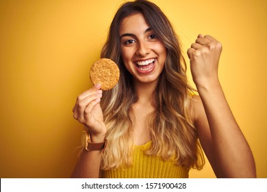 Young beautiful woman eating biscuit over grey isolated background screaming proud and celebrating victory and success very excited, cheering emotion