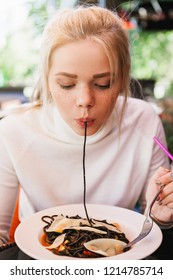 Young Beautiful Woman Eating Black Pasta With Seafood And Cuttlefish Ink In The Outdoor Restaurant. Funny And Beautiful.