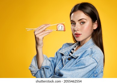 Young Beautiful Woman Eating Sushi, Holding Food With Chopsticks And Looking Sassy At Camera, Concept Of Asian Food Takeout, Yellow Background