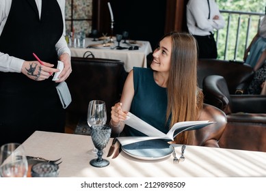 A Young Beautiful Woman In An Elegant Restaurant Looks Through The Menu And Makes An Order To A Young Waiter In A Stylish Apron. Customer Service In The Restaurant And Catering Establishments.