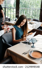 A Young Beautiful Woman In An Elegant Restaurant Looks Through The Menu And Makes An Order To A Young Waiter In A Stylish Apron. Customer Service In The Restaurant And Catering Establishments.