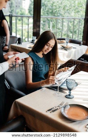 A young beautiful woman in an elegant restaurant looks through the menu and makes an order to a young waiter in a stylish apron. Customer service in the restaurant and catering establishments.