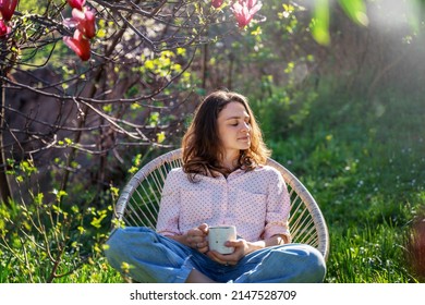 Young beautiful woman enjoying relaxing in the spring while sitting in the blooming garden of her home with a cup of tea