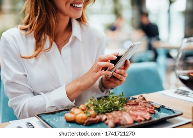 Young Beautiful Woman Enjoying In Tasty And Nicely Decorated Meal. She Sitting In Expensive Restaurant And Using Her Smart Phone For Food Photography.