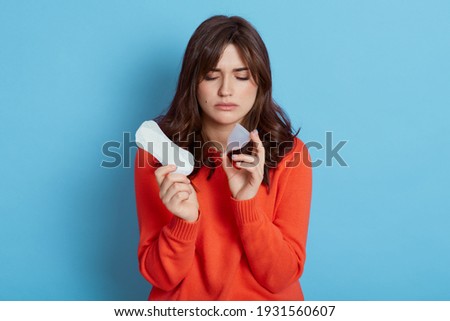 Young beautiful woman European young woman wearing orange sweater making choice between menstrual cup and hygiene pad, suffering from pain, need to choose best hygienic product, isolated on blue wall.
