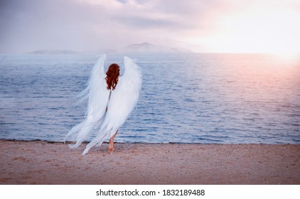 Young beautiful woman fallen angel stands on sea beach enjoy nature. costume bird white wings. Silhouette back view of mysterious red-haired girl at sunset. Bright divine sunlight, blue water ocean