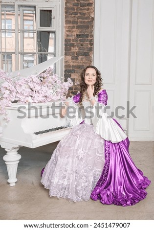 Young beautiful woman in fantasy white and purple rococo style medieval dress sitting near piano with pink flowers