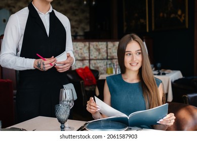 A young beautiful woman in a fine restaurant looks at the menu and makes an order to a young waiter in a stylish apron. Customer service.
