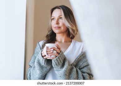 Young beautiful woman forty year with blonde long curly hair in cozy knitted grey sweater with cup of tea in hands in bright interior at home - Shutterstock ID 2137200007