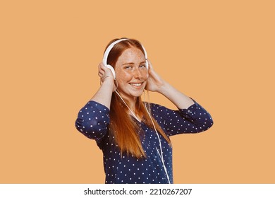 Young Beautiful Woman With Ginger Hair Red Head, Freckles And Blue Eyes, Wears Headphones Listening To Music And Dancing At Studio Over Beige Background. Technology Concept.