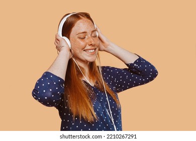 Young Beautiful Woman With Ginger Hair Red Head, Freckles And Blue Eyes, Wears Earphones, Listens Music With Eyes Closed At Studio Over Beige Background. Technology Concept.