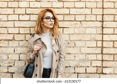 Young beautiful woman in glasses and a coat stands near a brick wall and looks aside