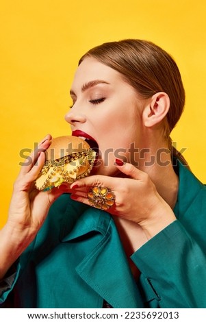 Young beautiful woman in green coat eating, biting cheeseburger with necklaces over yellow background. Luxury food. Food pop art photography. Complementary colors. Copy space for ad, text
