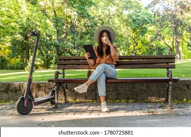 A young beautiful woman with a hat is sitting on a park bench with the tablet in her hands while drinking coffee. An electric scooter is parked next to her, while trees predominate in the background. - Powered by Shutterstock