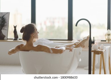 Young beautiful woman having bubble bath and looking at window. Back view.