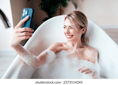 Young beautiful woman having fun while lying in bathtub full of foam at home. Charming smiling model relaxing in luxury bath interior. Female holding smartphone, looking at cellphone screen, use apps - Powered by Shutterstock