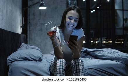 Young beautiful woman having late night video call, using her smartphone, sitting on bed, drinking wine. Long distance relationship concept