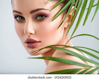 Young Beautiful Woman With Healthy Skin Of Face And Palm Leaves. Closeup Fresh Face Of An Attractive Caucasian Girl With Green Plants. Model With Bright Brown Eye Makeup. Skin Care Concept. 