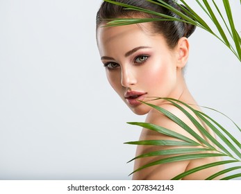 Young Beautiful Woman With Healthy Skin Of Face And Palm Leaves. Closeup Fresh Face Of An Attractive Caucasian Girl With Green Plants. Model With Bright Brown Eye Makeup. Skin Care Concept. 